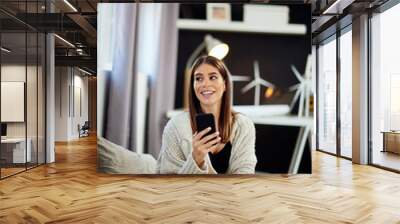 Beautiful smiling caucasian brunette sitting dressed in beige sweater sitting on bed in bedroom and using smart phone. Wall mural