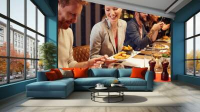 Handsome caucasian ginger taking food out of his girlfriend's plate while sitting in restaurant for dinner. In background are their friends. Wall mural