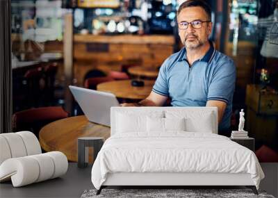 Mature entrepreneur sitting in cafeteria, using laptop for work and looking at camera. Wall mural