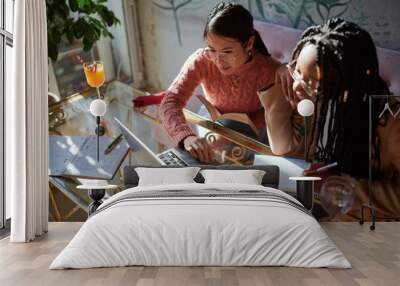 Two smart college girls sit in a coffee shop with books on a table and use a laptop for an online class. Girls on exchange study in a coffee shop. Wall mural