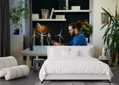 Young attractive bearded employee sitting in his modern office and using laptop for writing report about sustainability. Wall mural