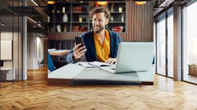 Young Caucasian businessman dressed smart casual using smart phone while sitting in cafe. On the desk laptop. Wall mural