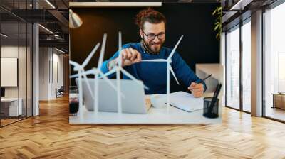 Young creative caucasian bearded environmentalist  with curly hair sitting in his office, touching windmill model and drawing new better model in notebook. Wall mural