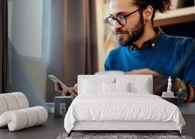 Young handsome bearded hipster sitting at his home office, holding cup of coffee and using smart phone. Wall mural