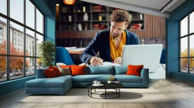 young serious bearded caucasian blogger dressed smart casual writing notes in agenda while sitting i Wall mural