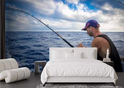 Tough man wearing baseball cap on board ship while fishing against blue sea background. Wall mural