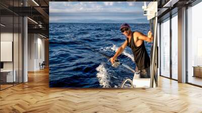 Tough man wearing baseball cap on board ship while fishing against blue sea background. Wall mural