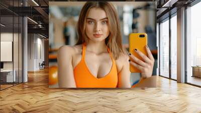 A fit girl in stylish activewear captures a fun gym selfie on her smartphone during a workout break. Wall mural