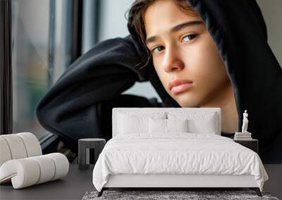 A lonely teen in a black hoodie sits by the window, absorbed in their phone, as the world outside remains distant. Wall mural