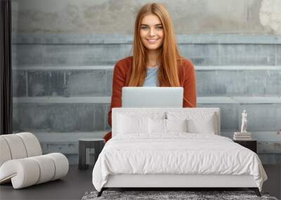 A stylish woman with long hair engrossed in her phone and laptop, seated on outdoor steps, enjoying her work. Wall mural