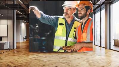 Senior male engineer training and explaining work to new employee wearing vest and safety jacket with hardhat helmet while pointing towards machine in factory and giving instructions Wall mural