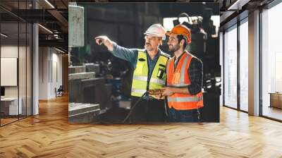 Senior male engineer training and explaining work to new employee wearing vest and safety jacket with hardhat helmet while pointing towards machine in factory and giving instructions Wall mural