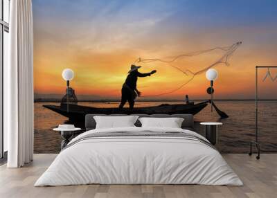 Silhouette of asian fisherman on wooden boat in action throwing a net for catching freshwater fish in nature river in the early morning before sunrise Wall mural