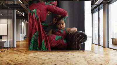 African mother dresses her daughter in traditional African cloth and turban, little girl sit on couch on dark background, mother-daughter relationship concept Wall mural