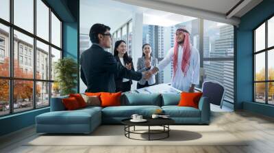 Arab businessman shaking hands with Asian businessman in the black suit near window, against building background, team applauded their congratulations for the business deal Wall mural