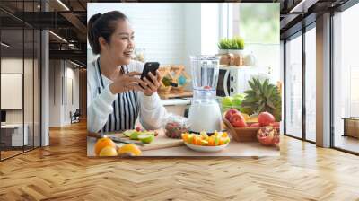 Asian middle age woman holding smart phone and looking out window, thinking something,  healthy foods are on the table in the kitchen Wall mural