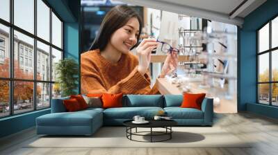 Beautiful girl choosing a glasses in optician store, Asian girl in sweater with different models of glasses Wall mural