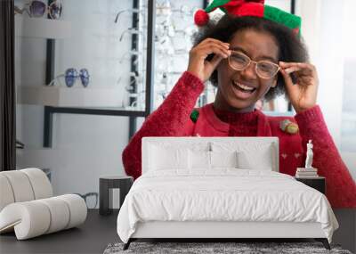 Dark skinned girl wears X'mas hat with new glasses in optical shop, young woman is choosing a glasses in optician store, showing her new glasses and smiling Wall mural