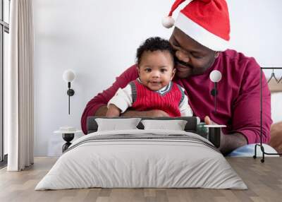 Happy African American father wears Santa hat holds gift box for his son on Christmas, little baby boy sit on fathers lap, winter holidays and people concept Wall mural