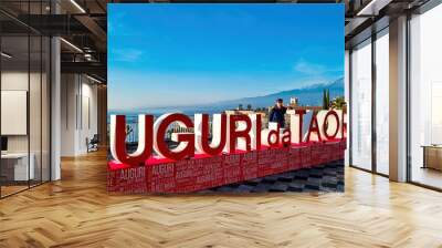 Man with christmas tree and signs of Christmas wishes (Auguri) on the main square (Piazza IX Aprile) in Taormina, Province of Messina, Sicily, Italy, Europe, EU. Holidays at the Mediterranean sea Wall mural