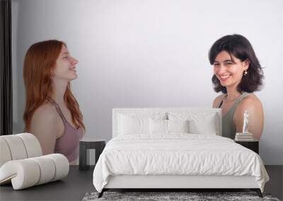 Two young women smiling and facing each other against a white background, expressing happiness and friendship. Wall mural