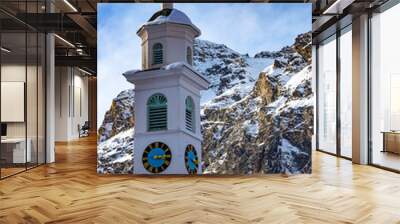 church tower with clock in mountain village, roof with snow, mountains background and blue sky Wall mural