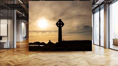 Silhouette of a Celtic cross with the evening sun directly behind it.  With a lighthouse, buildings and a cross in the background Wall mural