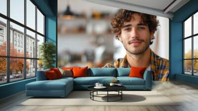 Young adult male with curly hair and a checkered shirt smiles gently, standing in a cozy kitchen with shelves in the background. Wall mural