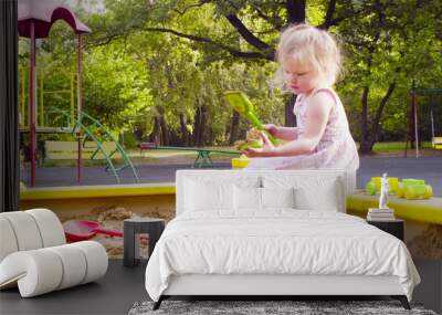 Low angle view. A little girl sitting in a sandbox is picking up sand with a scoop in a mold Wall mural