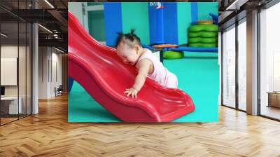 A cute Taiwanese baby girl is lying on the red slide.     Wall mural