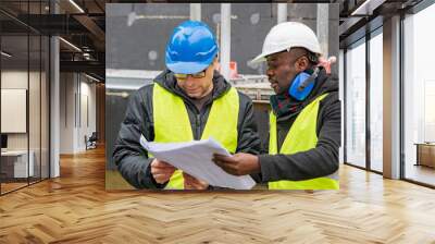 Civil engineers with hardhat and yellow jacket checking technical drawings and office blueprints among scaffolding on construction site Wall mural