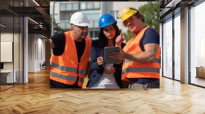 Senior foreman wearing safety vest, helmet and goggles providing instructions to a male and a female colleagues using a digital tablet computer on construction site Wall mural