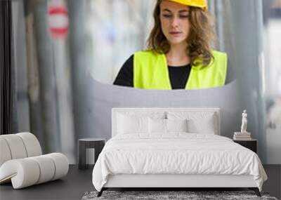 Vertical shot of cute female engineer with helmet and safety jacket on construction site examining office blueprints. Outdoors Wall mural