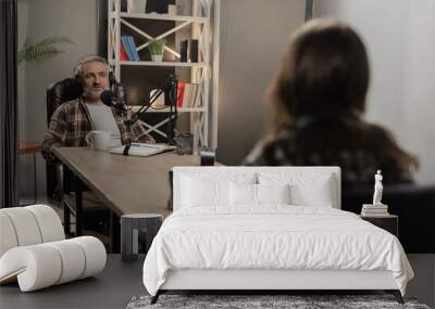 A man in headphones sits at a table opposite a woman and says something into a microphone. Two bloggers are recording a joint podcast from the studio. Wall mural