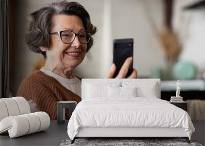 Happy mature middle aged woman enjoying using mobile apps, texting, sitting on sofa, smiling senior woman holding smart phone, looking at mobile phone screen, browsing social media at home Wall mural