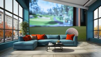 A golf ball is sitting on a green grassy field. The ball is white and has a small black dot on it. The field is surrounded by a green wall, and there is a golf simulator in the background Wall mural