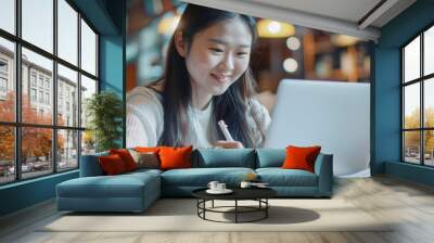 Euphoric Asian lady taking notes on laptop while studying and surfing the web at a casual cafe. Wall mural