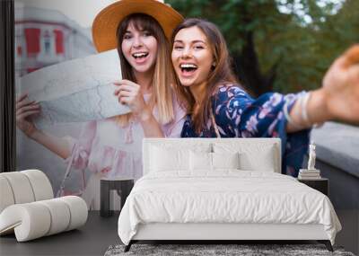 Happy travel together of two fashionable girls in sunny city centre. Young joyful women expressing positivity, using map, vacation with bags, camera, making photo, cheerful emotions, great mood Wall mural