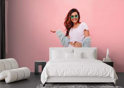 Laughing brunette woman in white t-shirt and mint coat looking and pointing away with thumb over pink background. funny surprised girl showing empty copy space with her hand Wall mural