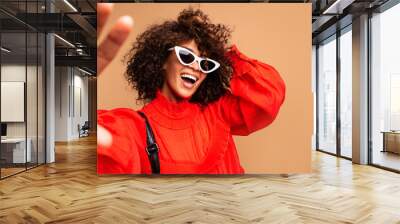 Portrait of beautiful young black woman taking selfie.Portrait of a pretty young afro american woman in retro style clothes smiling while standing and taking a selfie isolated over beige. Wall mural