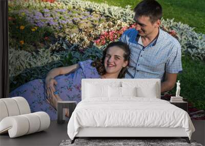 Young happy family, husband and pregnant wife lie on the grass on a bedspread in a park Wall mural