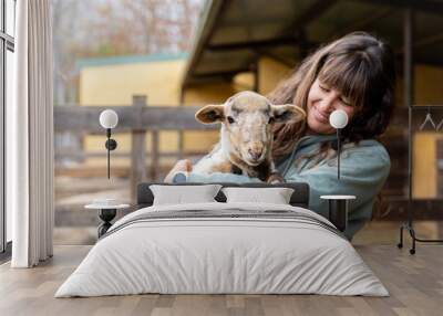 Happy young farmer woman hugging a baby sheep on a rural organic farm. Animal welfare and care in a barn. Wall mural