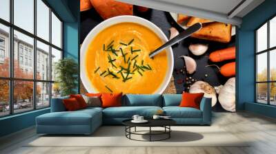 Top view to bowl of sweet potato cream soup with fresh chive and spoon, pepper, carrot, garlic and roasted toast bread on slate slab. Wall mural