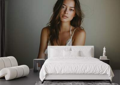 A beautiful woman with long brown hair, wearing a white lace top, sits against a pale grey wall. She looks directly at the camera, her eyes are a deep brown. Wall mural