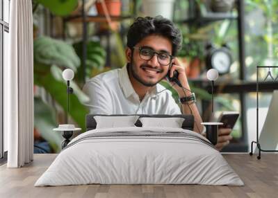 Smiling young Indian man freelancer talking on smartphone seated at desk with laptop, working at home, having business call, make phonecall to customer services or client, Generative AI Wall mural