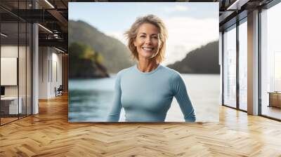 Portrait of a smiling woman in her 50s showing off a lightweight base layer against a beautiful lagoon background. AI Generation Wall mural