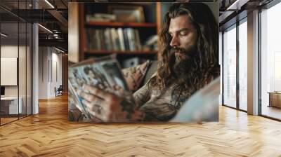 A bearded man with tattoos reads a magazine on a couch, surrounded by books. The cozy scene conveys a love for reading and relaxation. Wall mural