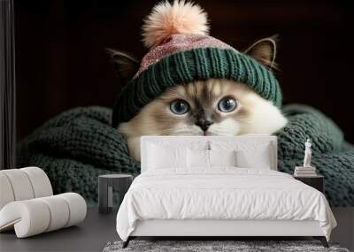 A white fluffy cat wearing a cozy green beanie with a pink pom pom rests its paws on a soft green knitted blanket. Wall mural