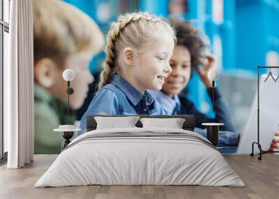 Group of three elementary students sitting at desk in school library and browsing Internet on laptop; focus on smiling blonde girl using touchpad Wall mural