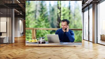 Middle aged businessman talking on cell phone and typing on laptop computer sitting at wooden table on country house terrace. Man working remotely in nature Wall mural